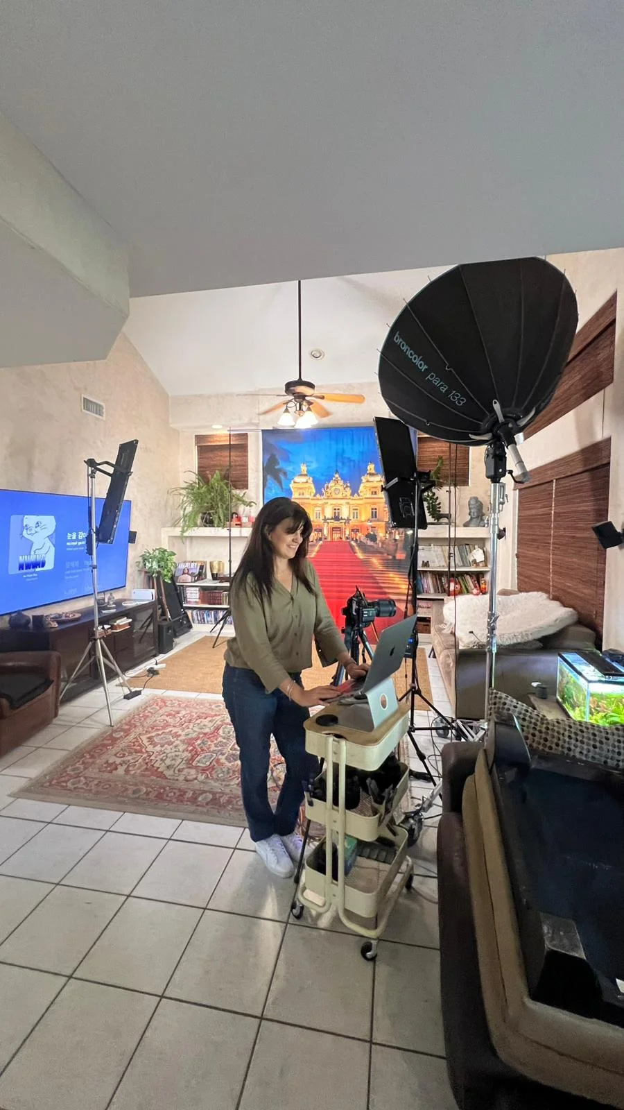 Behind the scenes of a professional backdrop photography setup showing photographer Marie working at her laptop with the Monte Carlo Casino backdrop visible in background, large Broncolor Para 133 softbox on right side, additional lighting equipment positioned around a living room studio space