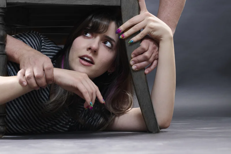 Young woman with an unusual theatrical expression looking up through a wooden chair frame with hands gripping the legs, showing the unpredictable human moments AI cannot replicate