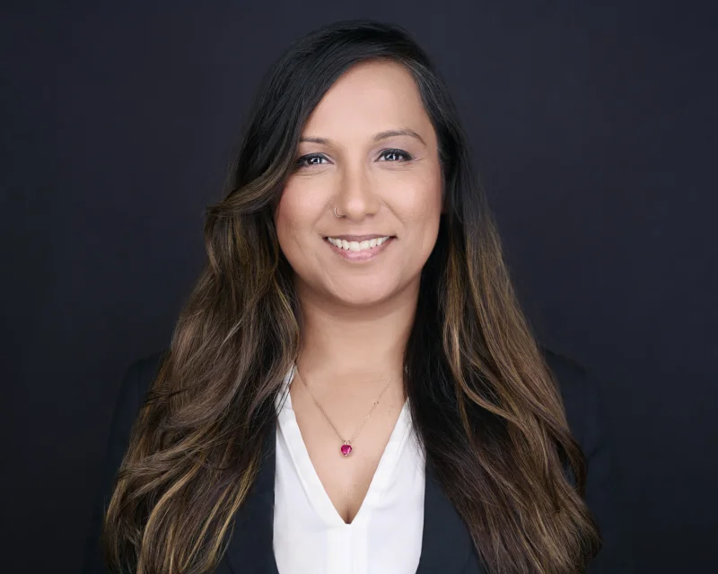 Woman in a black blazer over a white V-neck top smiling for a professional business headshot against a dark background