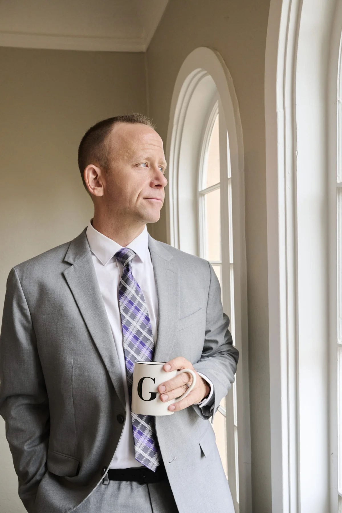 Attorney personal branding photography session in a gray suit with coffee by an arched office window Phoenix Arizona