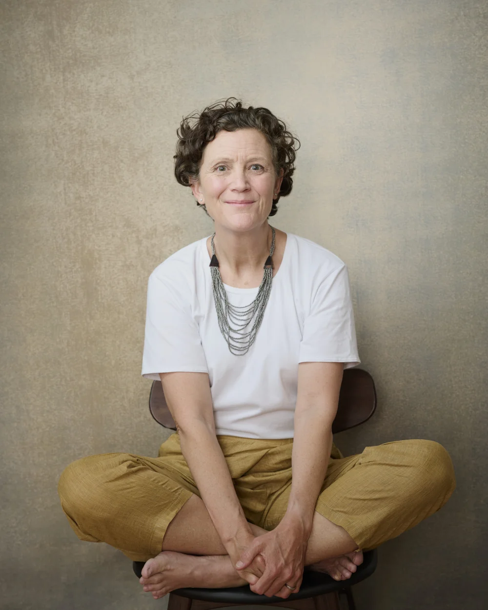 Lora Hebert, relaxed portrait seated cross-legged in white tee against warm textured backdrop by Marie Feutrier