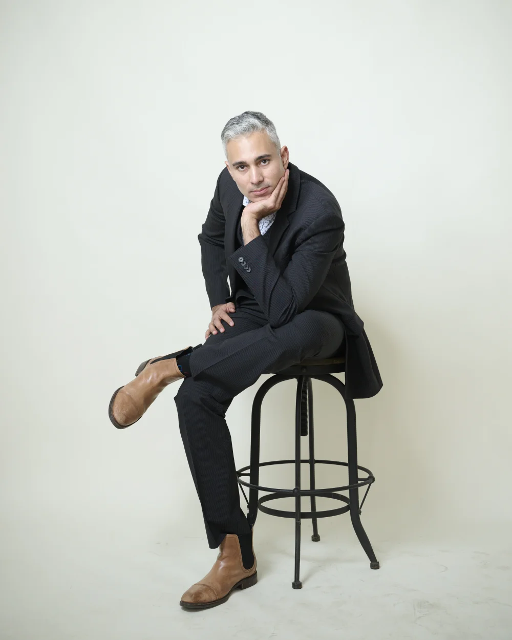 Rupesh Parbhoo, editorial portrait seated on stool in dark suit against cream backdrop by Marie Feutrier