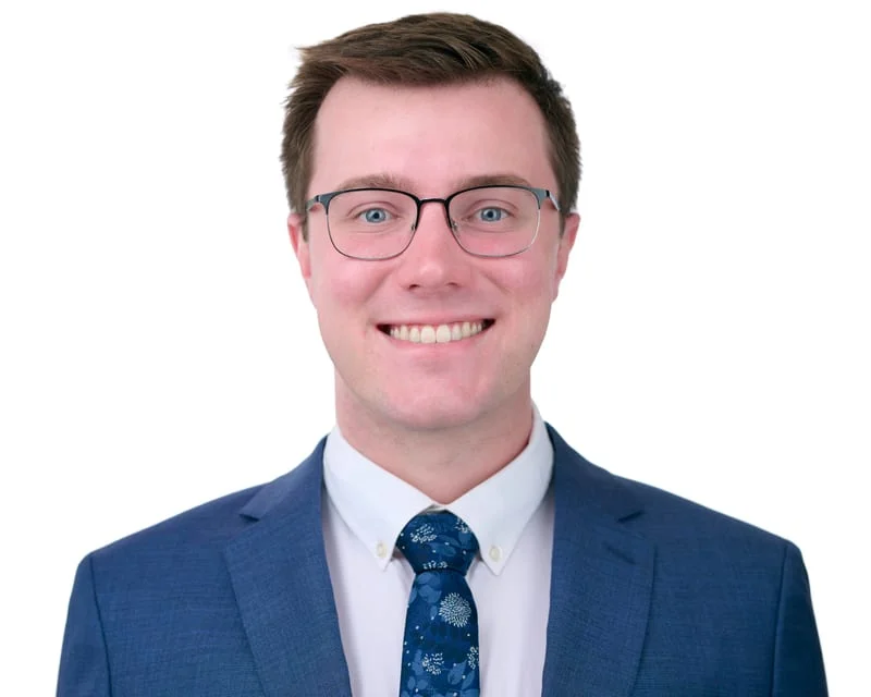 Male medical student in navy suit with blue floral tie and glasses smiling against white background ERAS wardrobe example